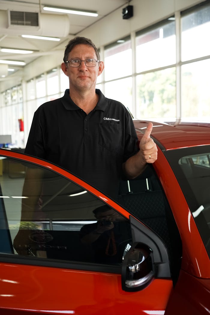 Smiling man giving thumbs up next to red car indoors, symbolizing satisfaction.